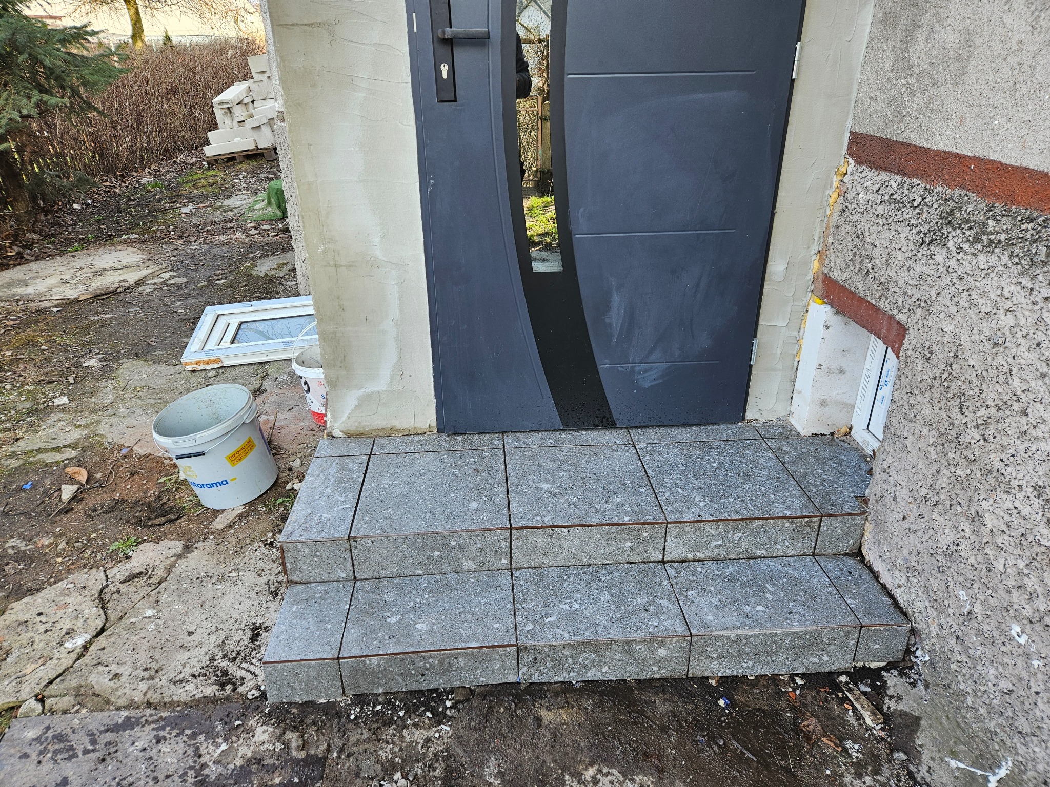 Ułożone płytki na schodach wejściowych do domu, widoczne drzwi wejściowe, wiadro z farbą i gruz. Prace wykończeniowe na zewnątrz budynku.