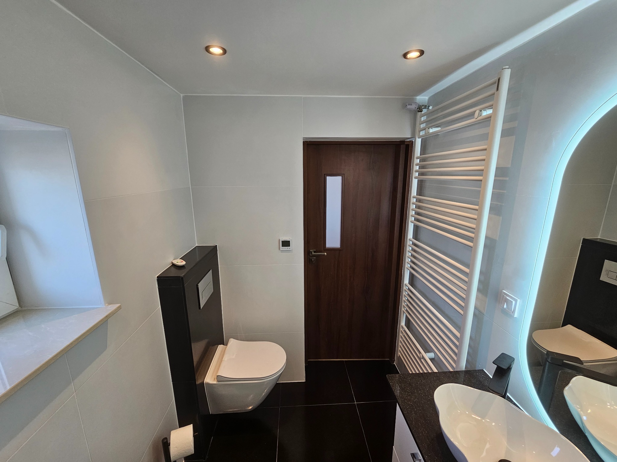 Nowoczesna łazienka z podwieszaną toaletą, czarną armaturą i grzejnikiem drabinkowym. Umywalka wpuszczana w czarny blat, lustro z podświetleniem LED.