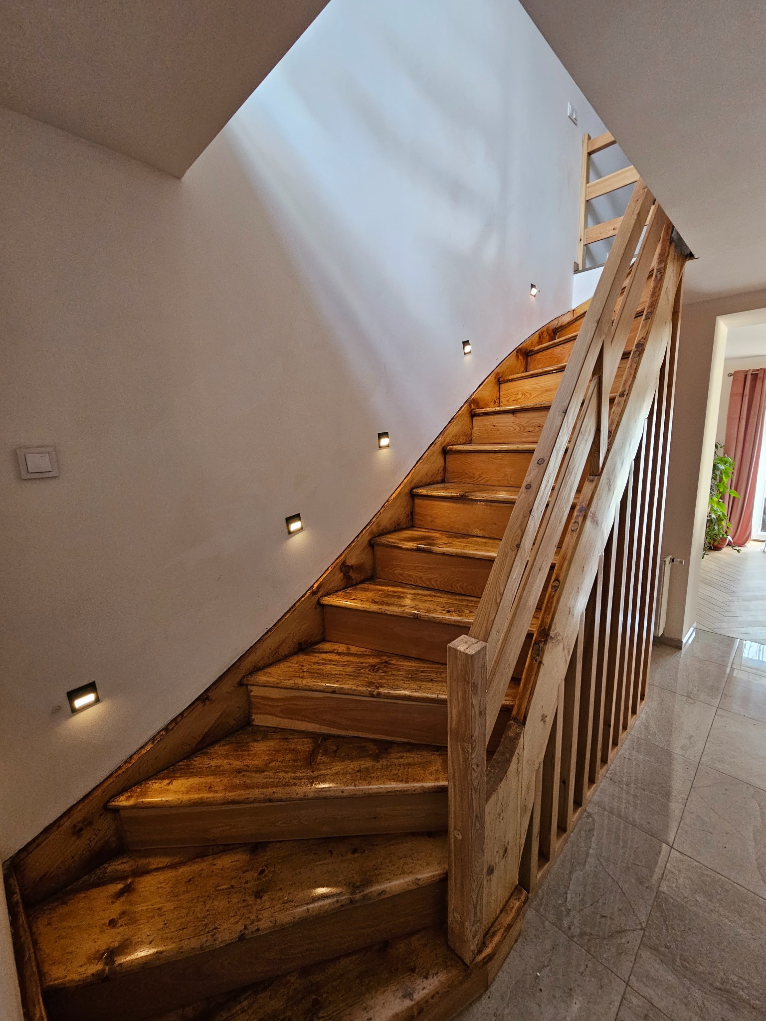 Drewniane schody z oświetleniem punktowym na ścianie, widok z dołu, ukazujący detale wykończenia i naturalne usłojenie drewna. Widoczny fragment podłogi z płytek.