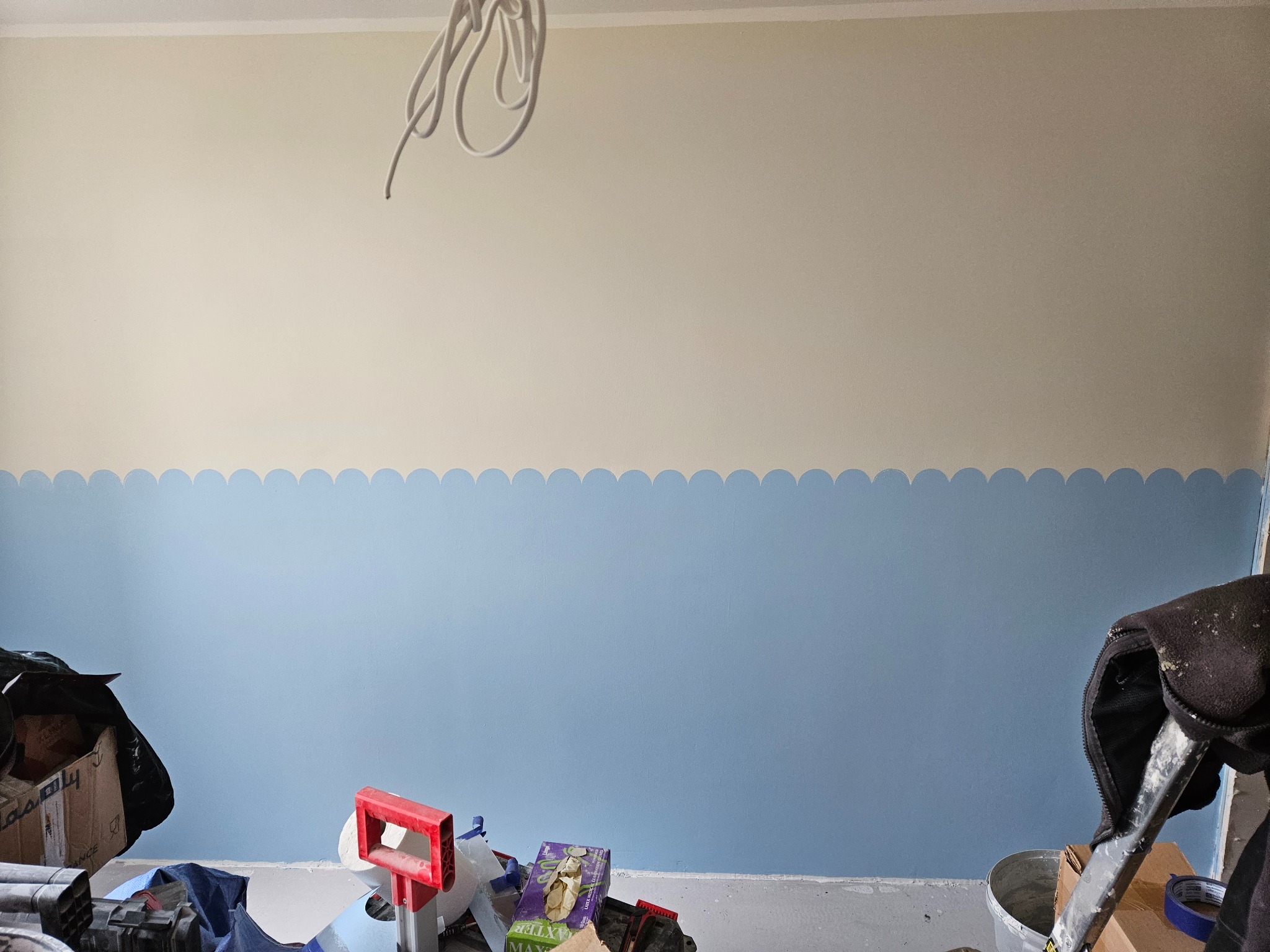 Ściana pomalowana na dwa kolory z dekoracyjnym wzorem falistym. Na podłodze widoczne narzędzia i akcesoria malarskie, sugerujące etap prac wykończeniowych.