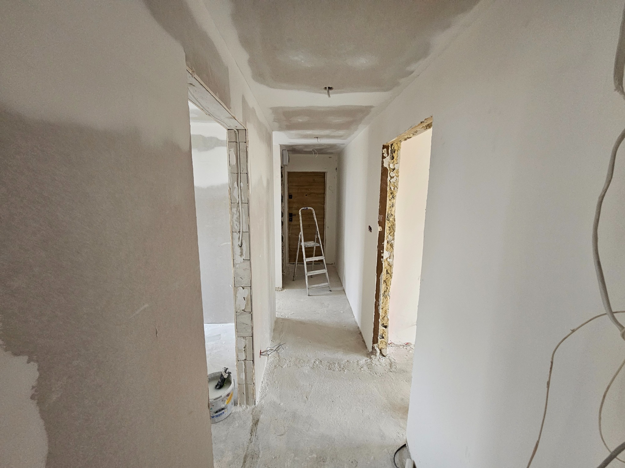 Korytarz w trakcie remontu: ściany i sufit po szpachlowaniu, widoczne ościeżnice drzwiowe, drabina w głębi. Przy ścianie stoi wiadro z zaprawą.
