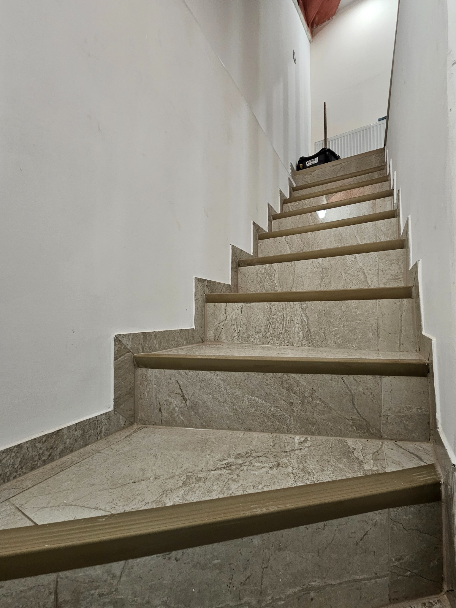 Wykończone schody z szarych płytek imitujących marmur, z brązowymi listwami antypoślizgowymi. Widok z dołu, na górze widoczny sprzęt budowlany.