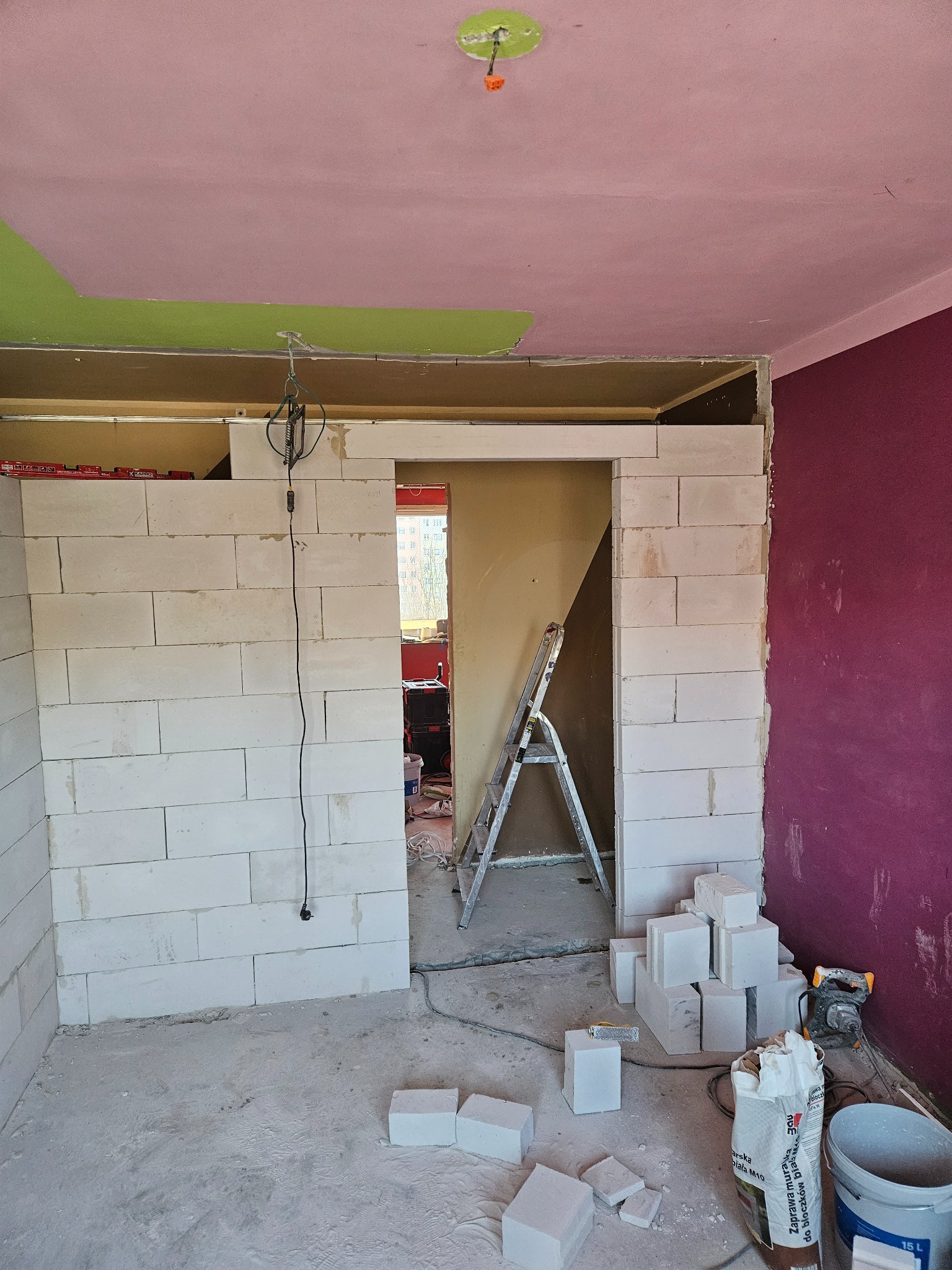 Wnętrze w trakcie budowy ściany z białych bloczków, widoczne narzędzia i materiały budowlane, sufit w kolorze różowym i zielonym, drabina w tle.