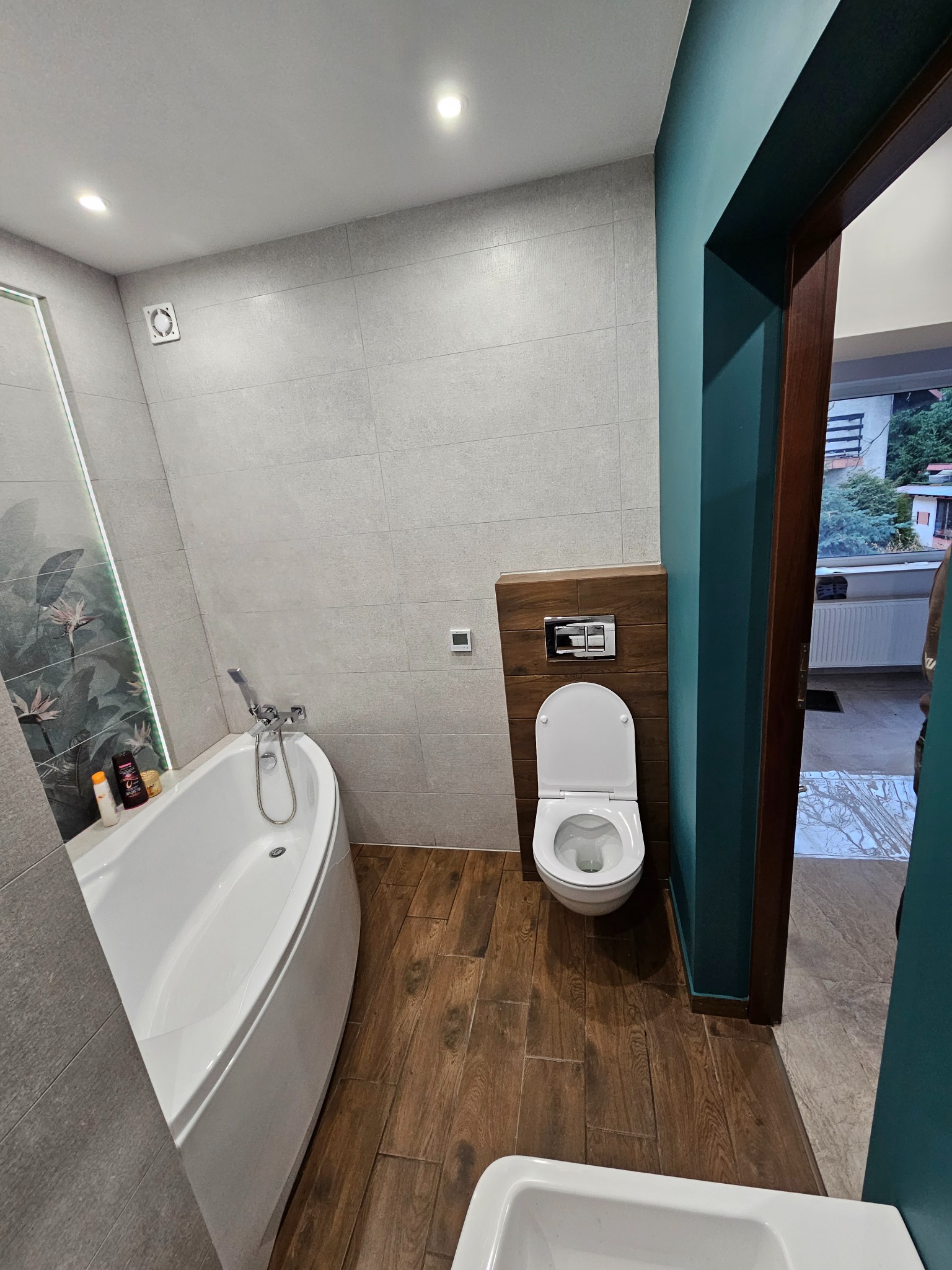 Wykończona łazienka w Lubominie z wanną, toaletą i drewnopodobną podłogą. Ściany wyłożone szarą płytką, akcent turkusowej farby przy drzwiach.