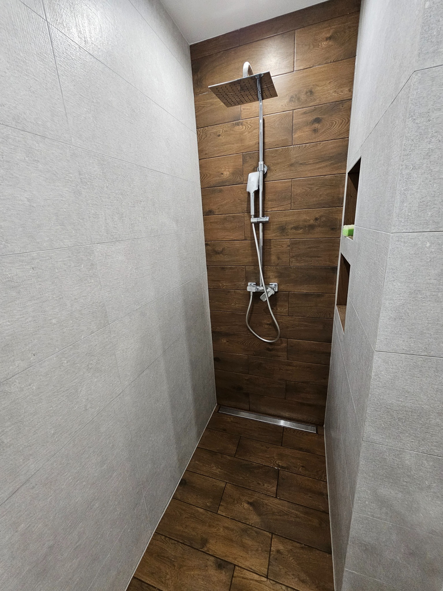 Nowoczesna kabina prysznicowa walk-in z drewnopodobnymi płytkami i szarymi ścianami. Widoczny odpływ liniowy i wnęka w ścianie.