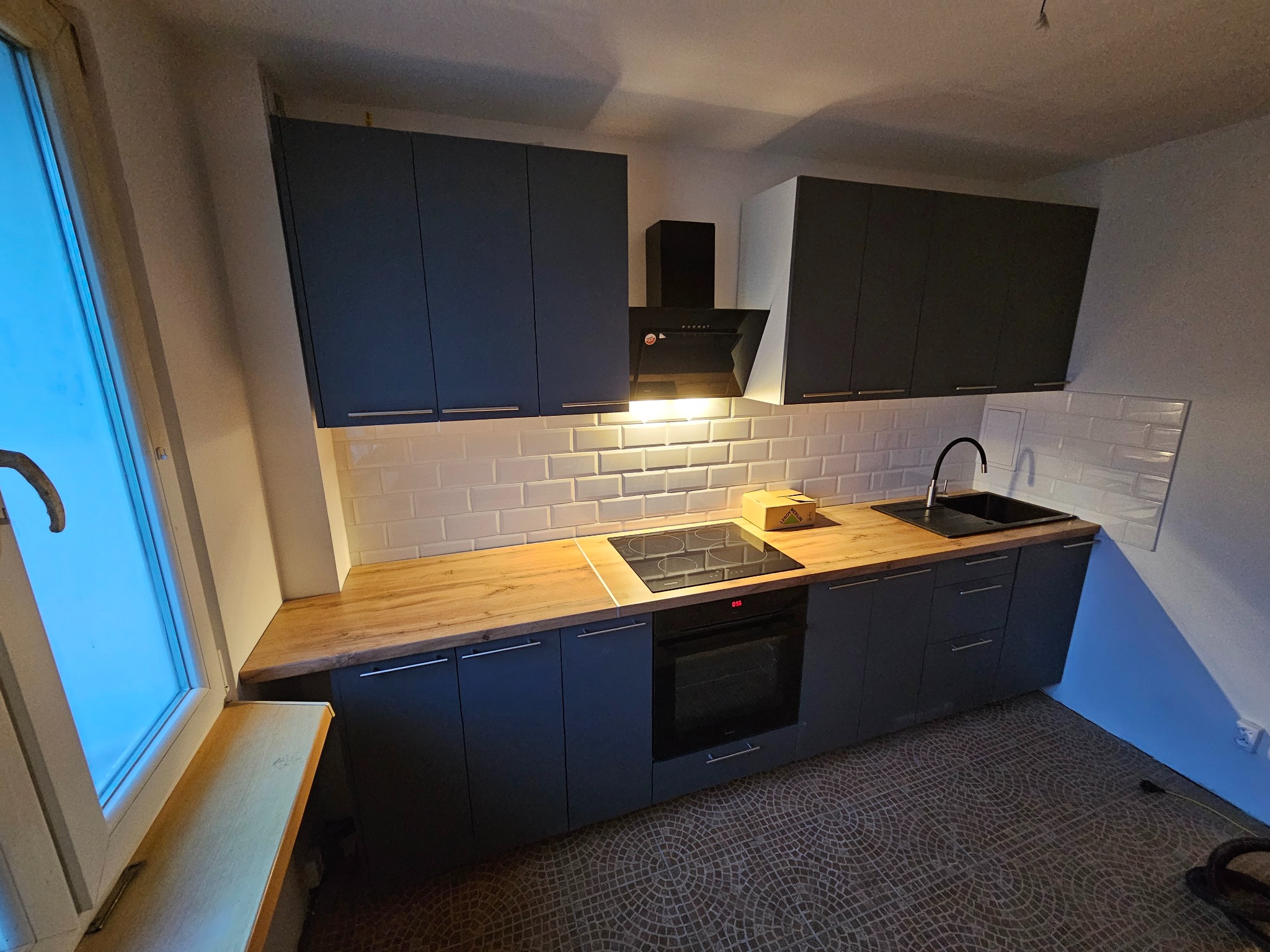 Nowoczesna kuchnia z szarymi szafkami, drewnianym blatem i białymi płytkami. Widoczny piekarnik, płyta indukcyjna i okap. Na podłodze mozaika.