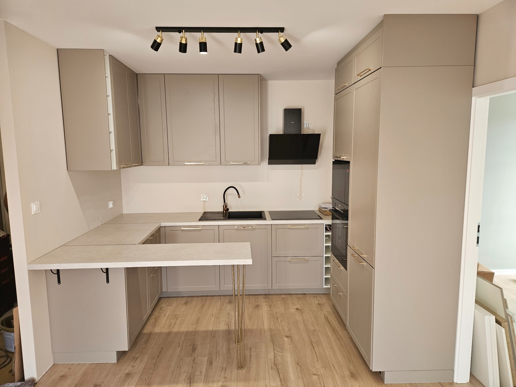 Jasna, nowoczesna kuchnia z szarymi szafkami i złotymi akcentami, oświetlona reflektorami. Widoczny fragment blatu z metalowymi nogami i sprzęt AGD.