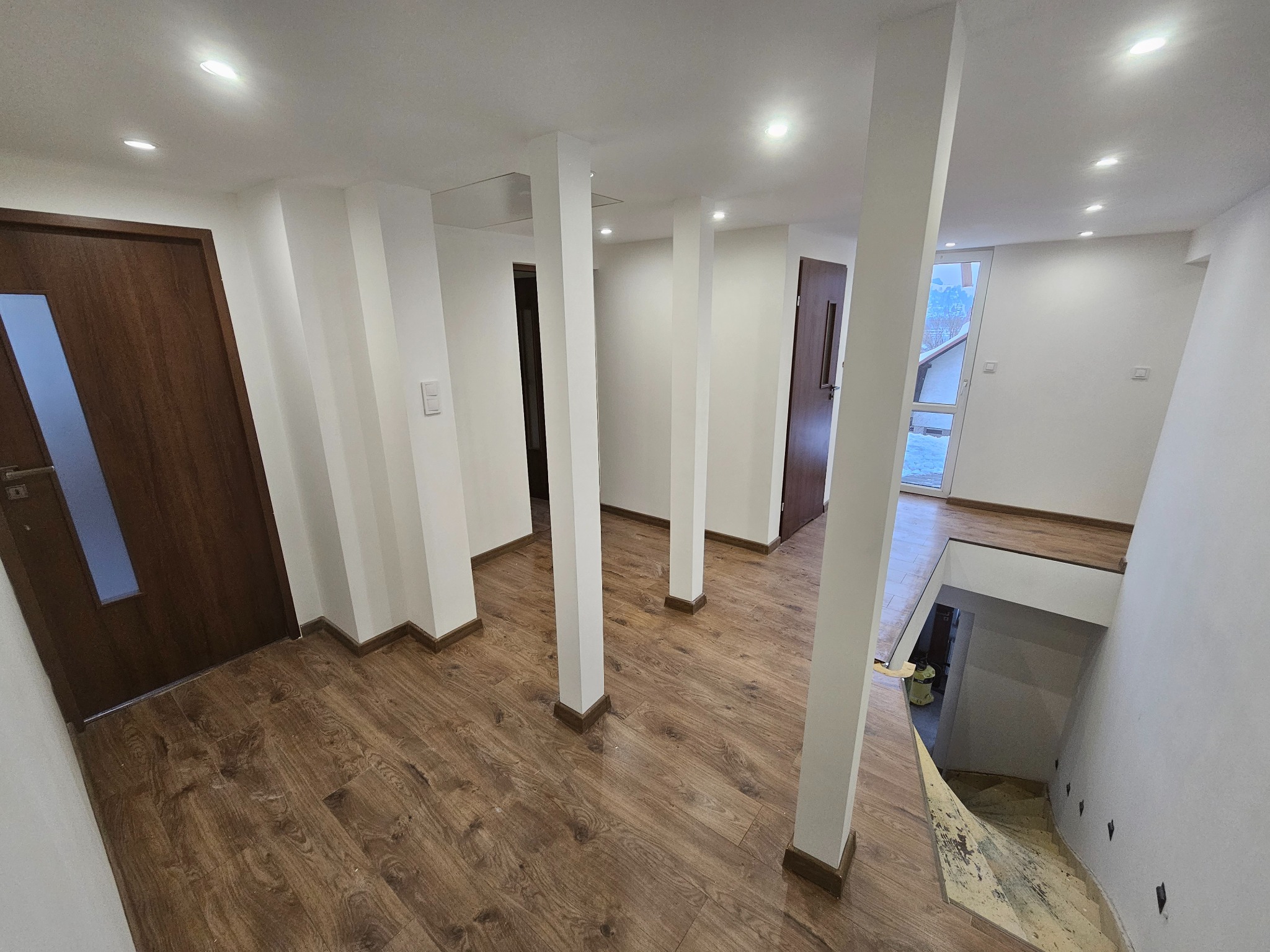 Jasne wnętrze z drewnianą podłogą i białymi kolumnami. Widoczne drzwi i schody w dół. Oświetlenie sufitowe.