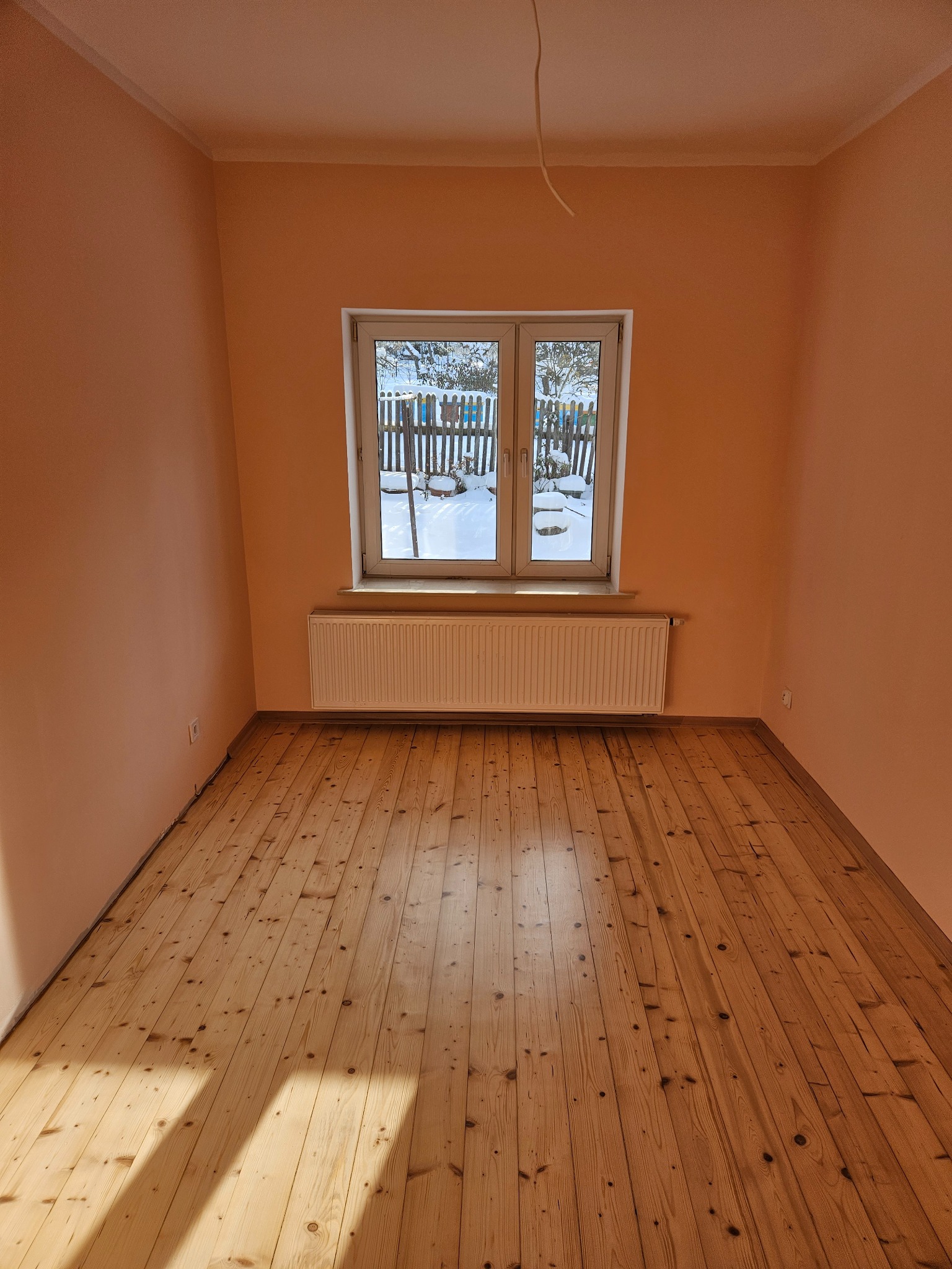 Pusty pokój po remoncie: świeżo pomalowane brzoskwiniowe ściany, odnowiona drewniana podłoga, białe okno z widokiem na zimowy ogród. Grzejnik pod oknem.