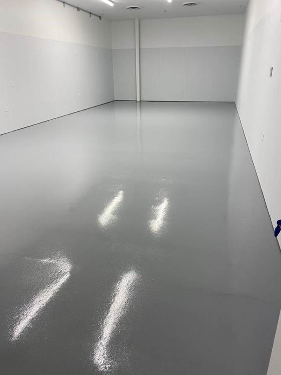 Świeżo wylana, gładka, szara podłoga w pustym pomieszczeniu przemysłowym z białymi ścianami i widocznymi śladami po czyszczeniu.