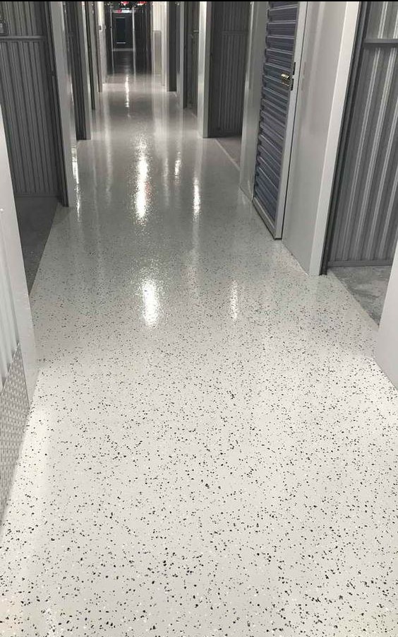 Biała posadzka w korytarzu magazynowym, wykończona drobnymi czarnymi plamkami, odbijająca światło sufitowe.