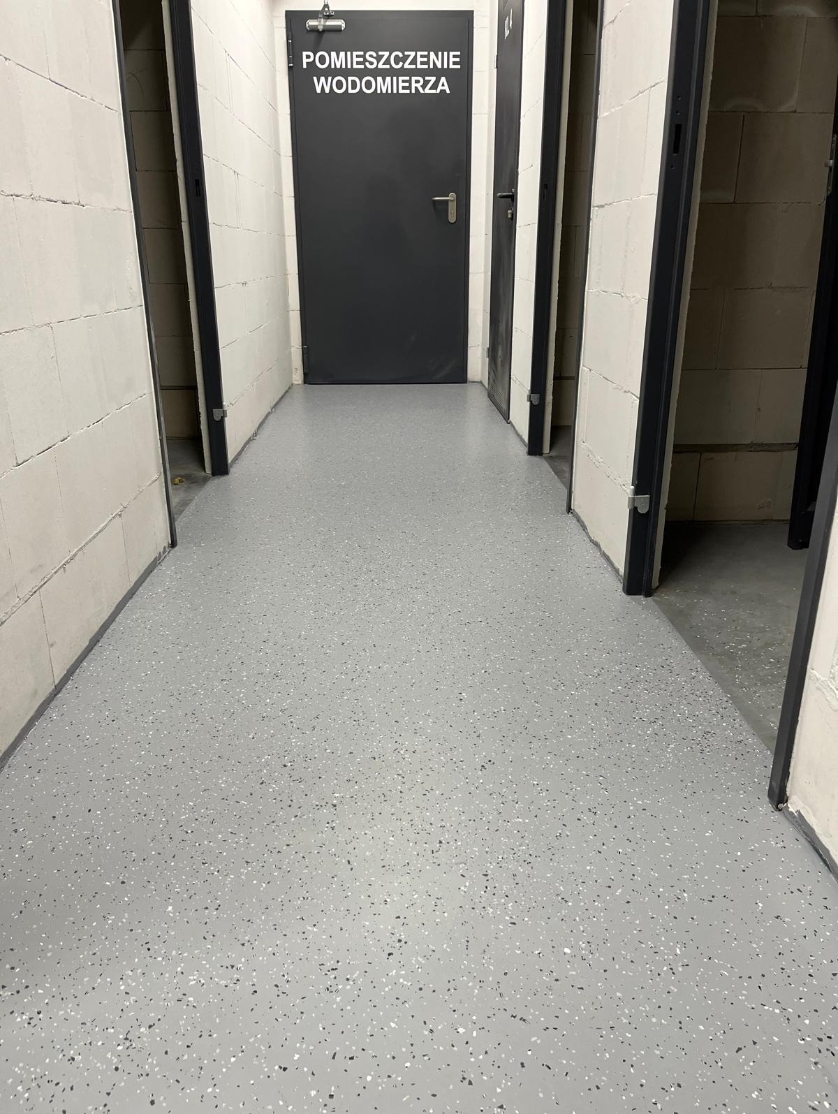 Szara, żywiczna posadzka w pomieszczeniu technicznym z drzwiami do pomieszczenia wodomierza.
