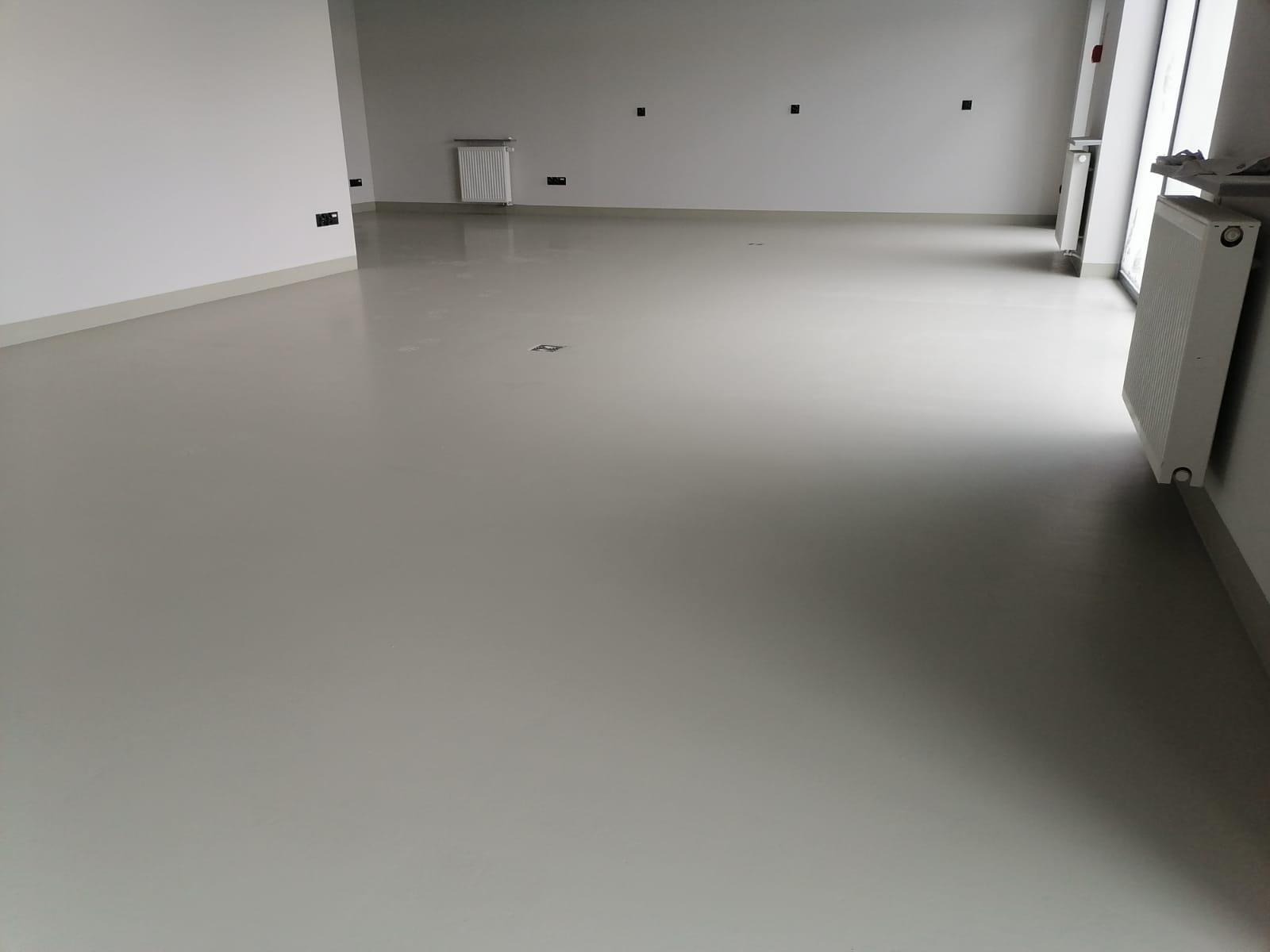 Jasnoszara, gładka podłoga w pustym pomieszczeniu z grzejnikami przy ścianach i gniazdkami elektrycznymi na ścianach.