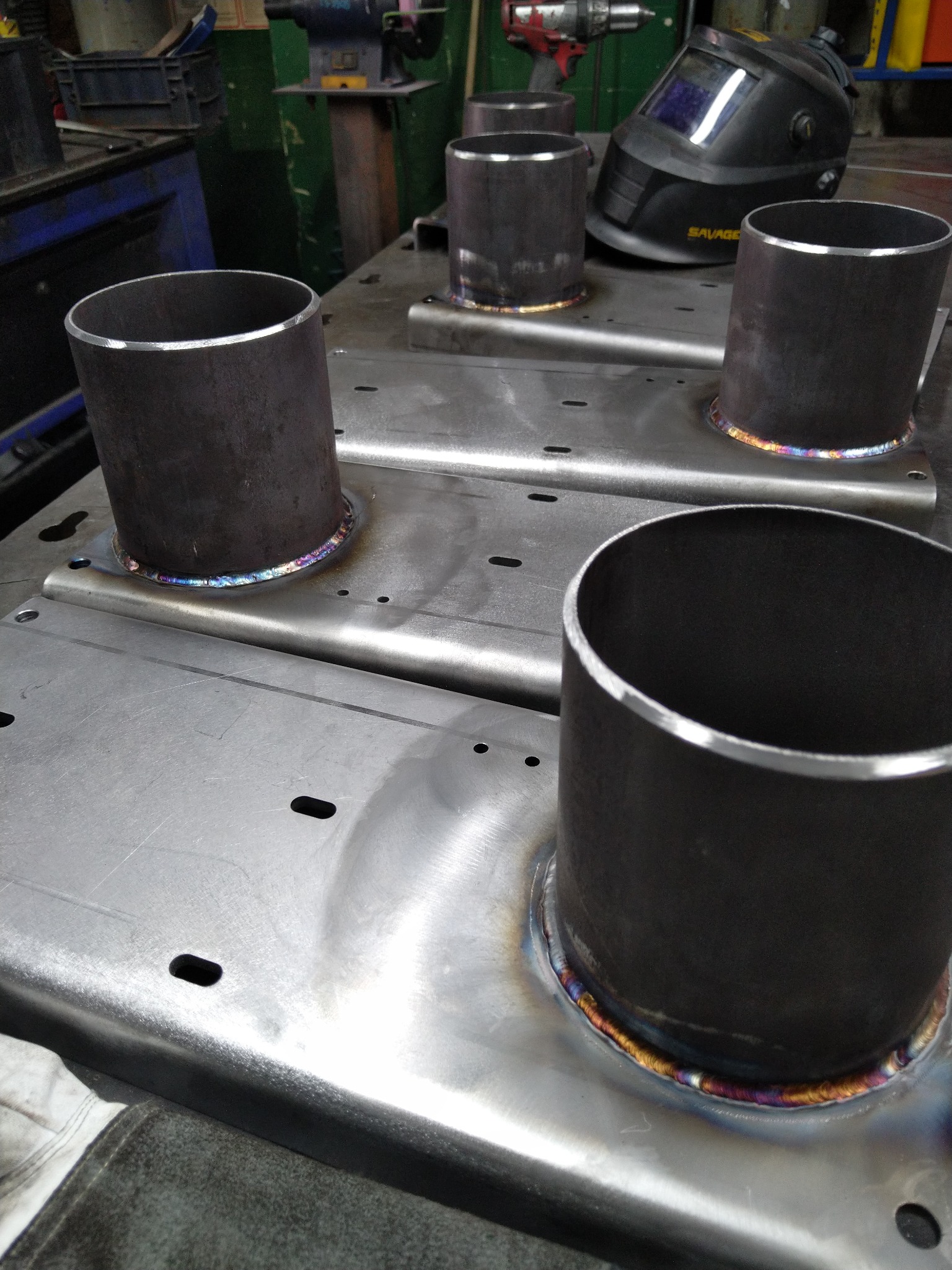 Metalowe rury przyspawane do blachy, widoczne tęczowe przebarwienia spoin, w tle maska spawalnicza i narzędzia warsztatowe.