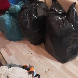 Firma Sprzątająca Blask - Worki na śmieci, w tym czarne i niebieskie, stoją na podłodze w pokoju obok torby z ubraniami, co sugeruje proces porządkowania lub opróżniania przestrzeni.
