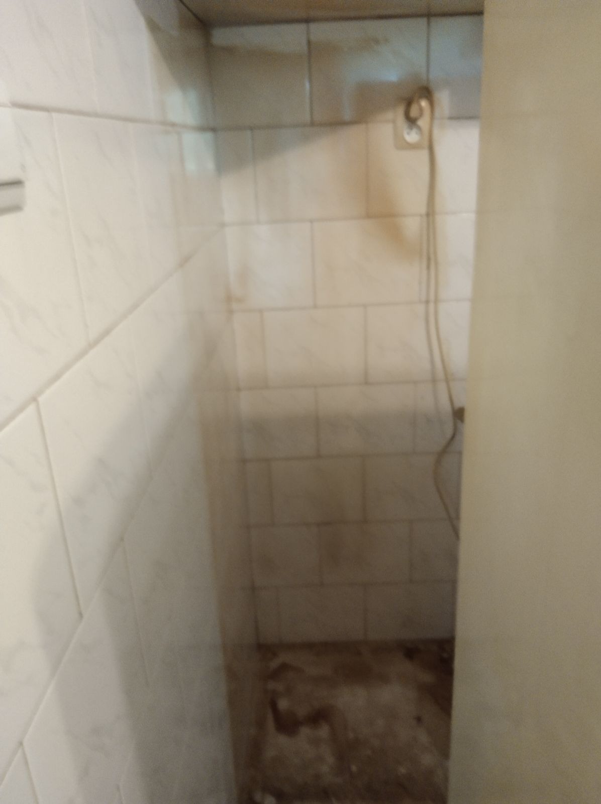 Zanieczyszczona, wykafelkowana wnęka z widocznym gniazdkiem elektrycznym i wiszącym kablem, wymagająca gruntownego wysprzątania w Gorzowie Wielkopolskim.