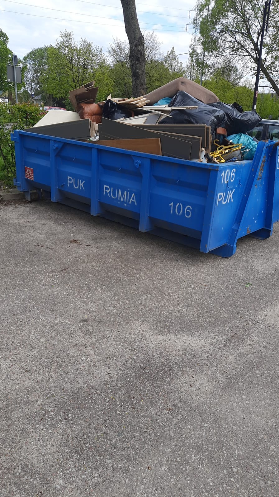 Niebieski kontener PUK Rumia wypełniony odpadami poremontowymi i meblami, widoczne płyty meblowe, fragment sofy i worki na śmieci.