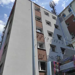 Wielopiętrowy budynek z fragmentami elewacji w trakcie prac, widoczne ocieplenie w paczkach, metalowe rury spustowe i uchwyty na elewacji, antena telefonii komórkowej na dachu sąsiedniego budynku.