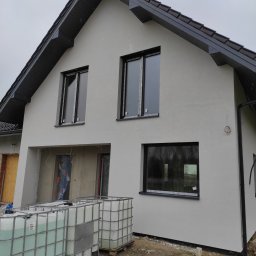 Ciepły Dom - Nowo otynkowany dom jednorodzinny z czarną stolarką okienną, widoczne zbiorniki z wodą na paletach przed budynkiem, stan surowy otwarty w części budynku.