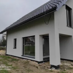 Nowoczesny dom jednorodzinny z szarą elewacją i czarną stolarką okienną, widoczne ślady prac wykończeniowych wokół budynku, pochmurne niebo.