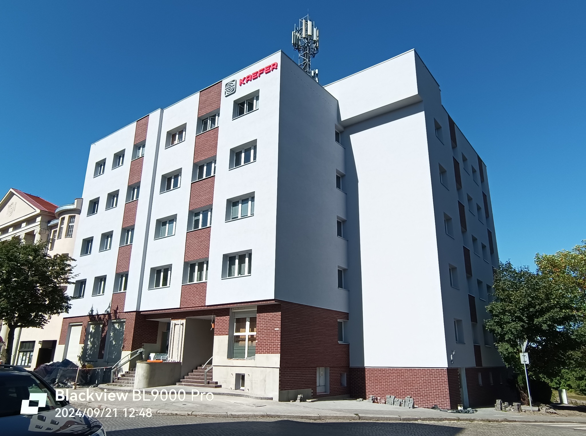 Wielopiętrowy budynek po renowacji elewacji, z białymi ścianami i elementami z czerwonej cegły, logo firmy na szczycie, antena telekomunikacyjna na dachu, niebo błękitne.