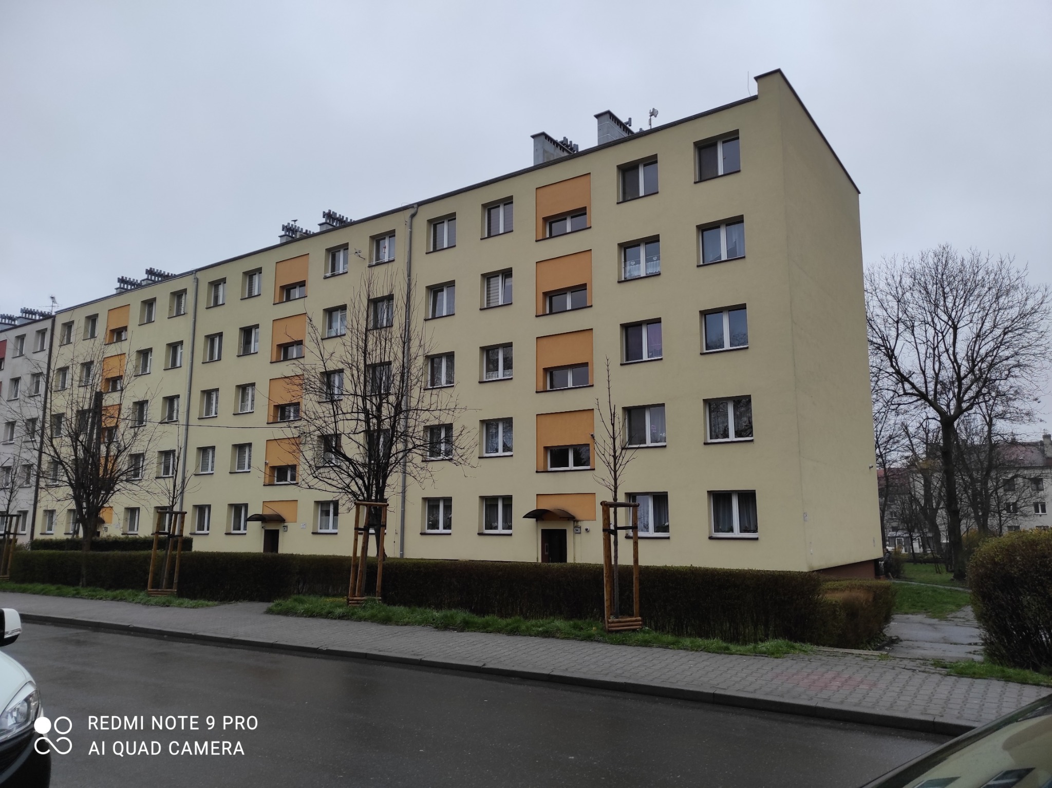 Wielopiętrowy budynek mieszkalny po renowacji elewacji, z elementami w kolorze beżowym i pomarańczowym, widoczny z poziomu ulicy w pochmurny dzień.