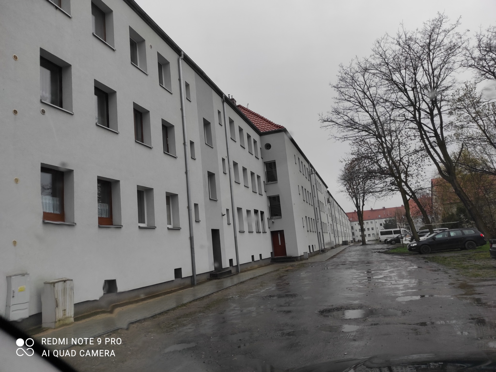 Długa, biała elewacja budynku wielorodzinnego z rzędami okien, widoczna z perspektywy mokrej ulicy w pochmurny dzień.