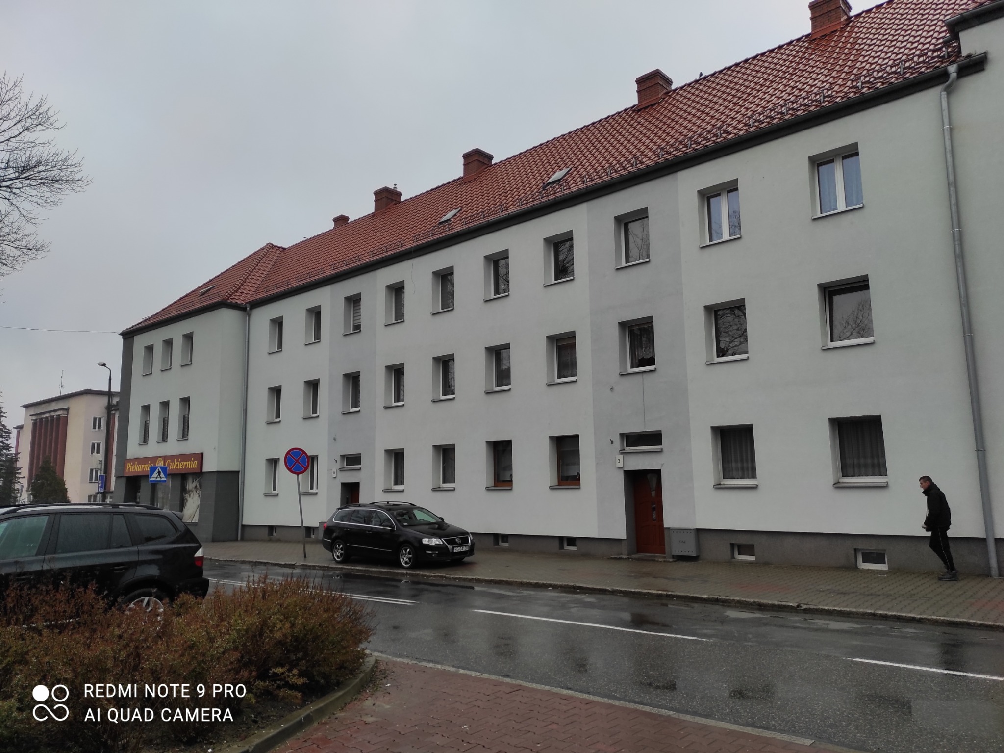 Szary budynek wielorodzinny z czerwoną dachówką, widok z ulicy w pochmurny dzień, zaparkowane samochody i przechodzień.