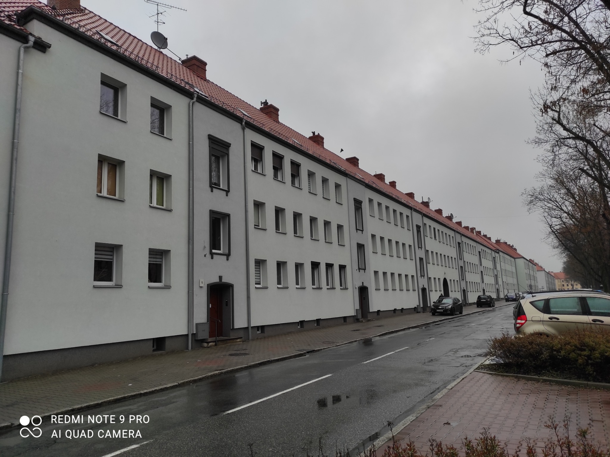 Długi szereg odnowionych, szarych budynków wielorodzinnych z czerwonymi dachami, widok z poziomu ulicy w pochmurny dzień, mokra nawierzchnia odbija światło.