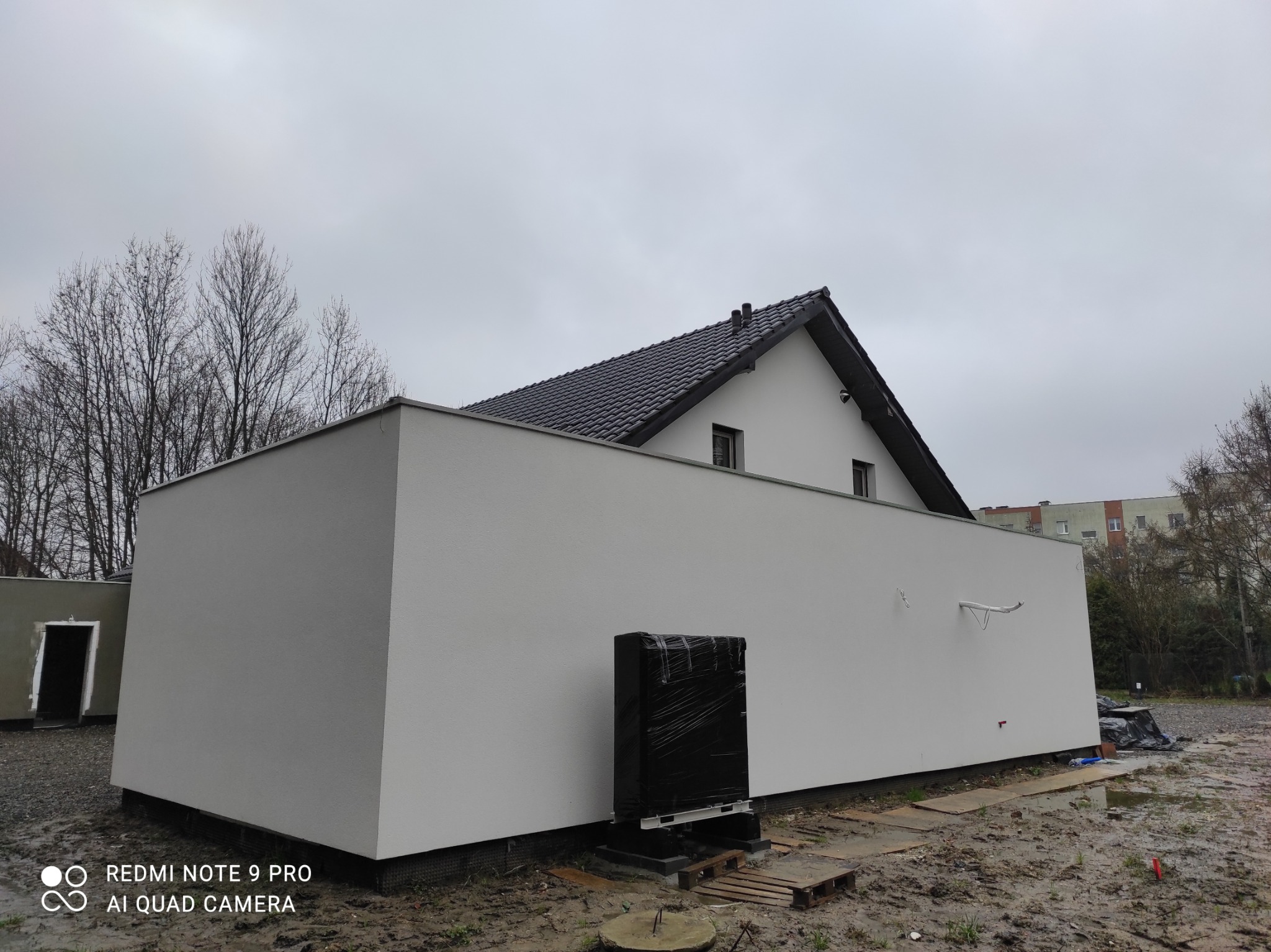 Nowoczesny budynek z jasną, gładką elewacją w Gliwicach, widok z zewnątrz, z czarnym dachem i elementami instalacji na ścianie, na wilgotnym terenie budowy.