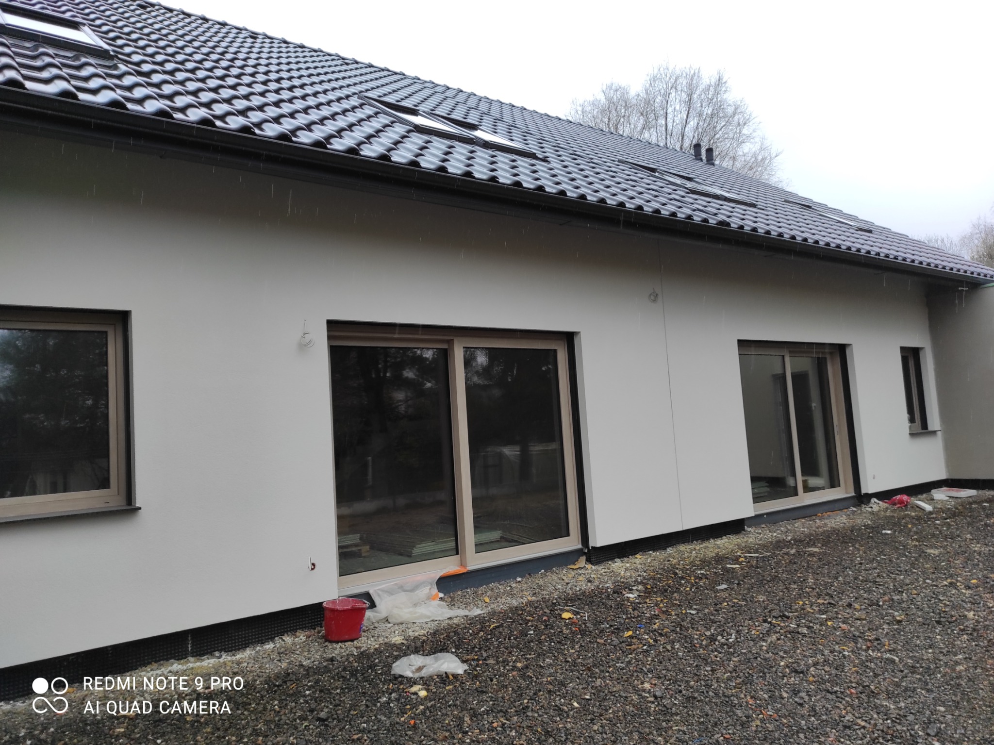 Nowoczesny dom jednorodzinny z jasną elewacją i grafitową dachówką, widoczne okna i drzwi przesuwne z jasnymi ramami, teren wokół domu wysypany żwirem, w tle drzewa bez liści.