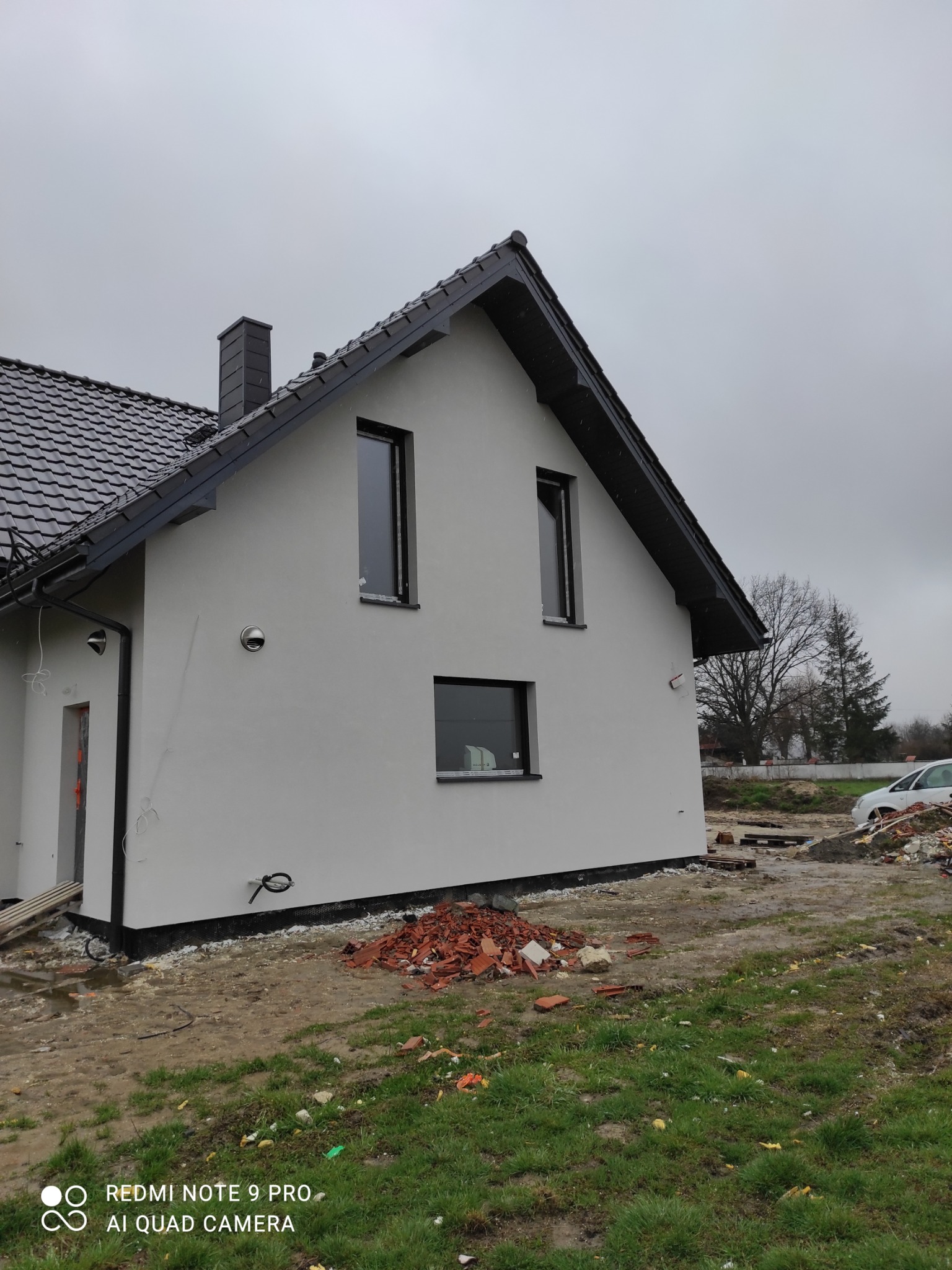 Nowoczesny dom jednorodzinny z jasnoszarą elewacją i ciemną dachówką, widok z boku, teren wokół domu w trakcie prac wykończeniowych, porozrzucane cegły.