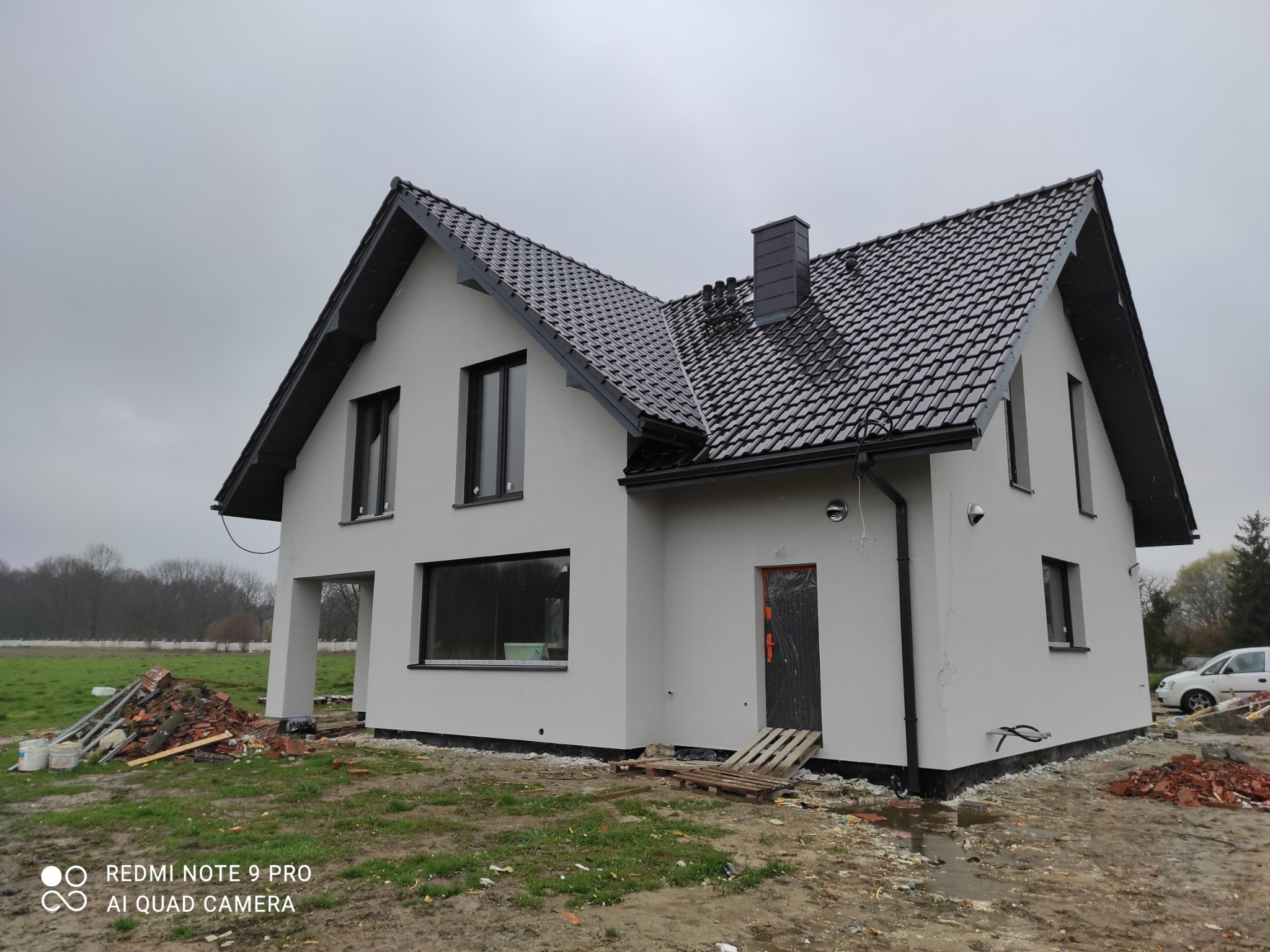 Nowoczesny dom jednorodzinny z szarą elewacją i czarną dachówką, w trakcie prac wykończeniowych na zewnątrz, widoczne materiały budowlane i palety, pochmurne niebo w tle.