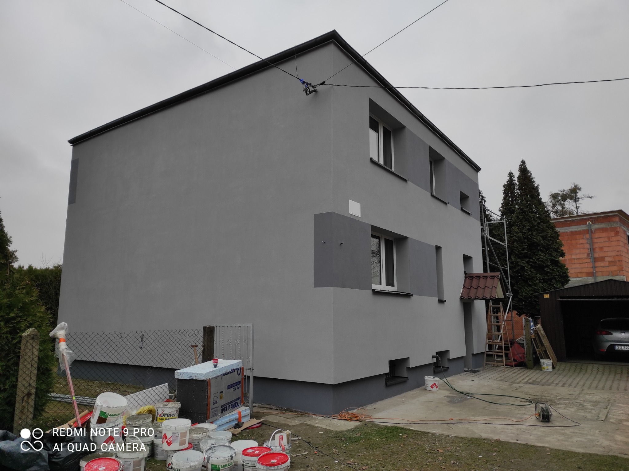 Dwukondygnacyjny dom jednorodzinny z szarą elewacją po remoncie, widoczne puszki po farbach, fragment rusztowania i garaż z samochodem.