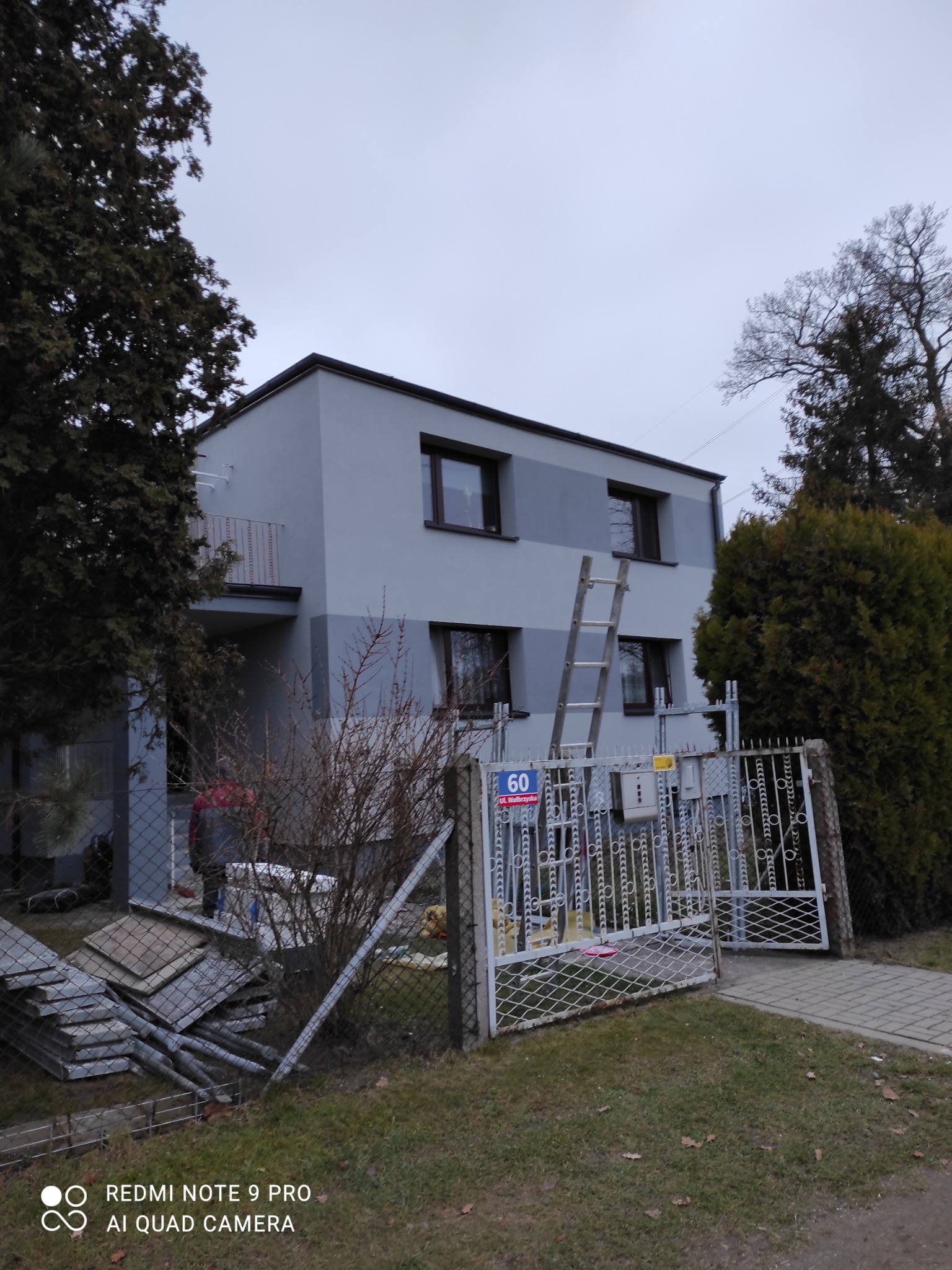 Dwukondygnacyjny dom z szarą elewacją, drabina oparta o ścianę, widoczny fragment ogrodzenia i sterta płyt izolacyjnych przed posesją.