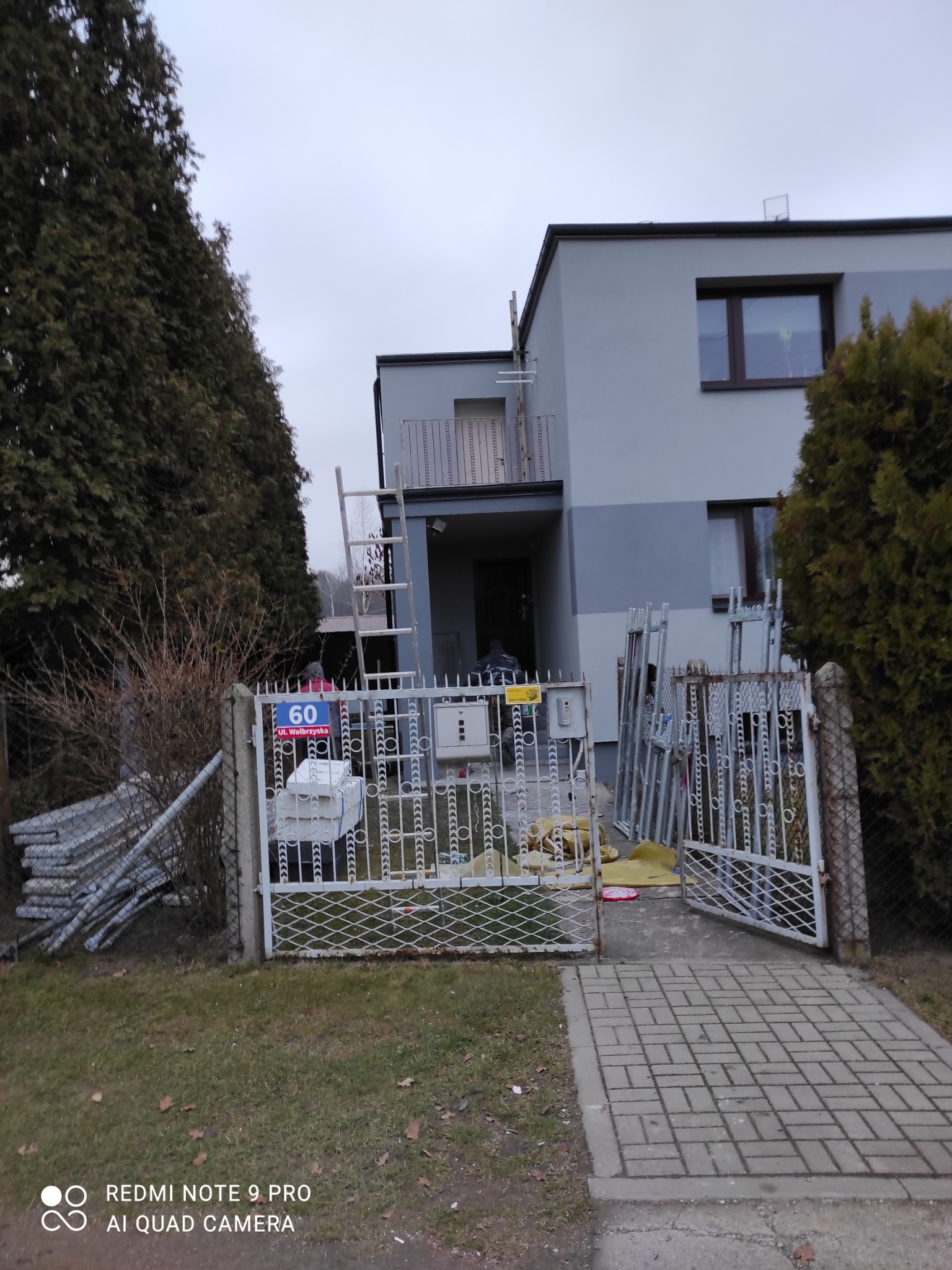 Dwukondygnacyjny dom jednorodzinny z szarą elewacją, widoczne rusztowania i drabina oparta o ścianę, przygotowania do prac elewacyjnych, brama wjazdowa z numerem posesji.