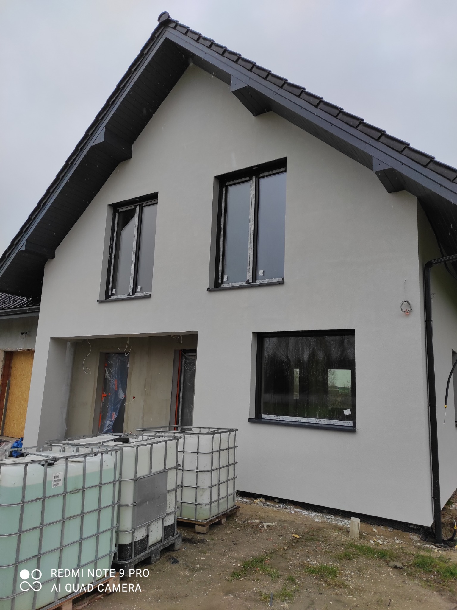Nowo otynkowany dom jednorodzinny z czarną stolarką okienną, widoczne zbiorniki z wodą na paletach przed budynkiem, stan surowy otwarty w części budynku.