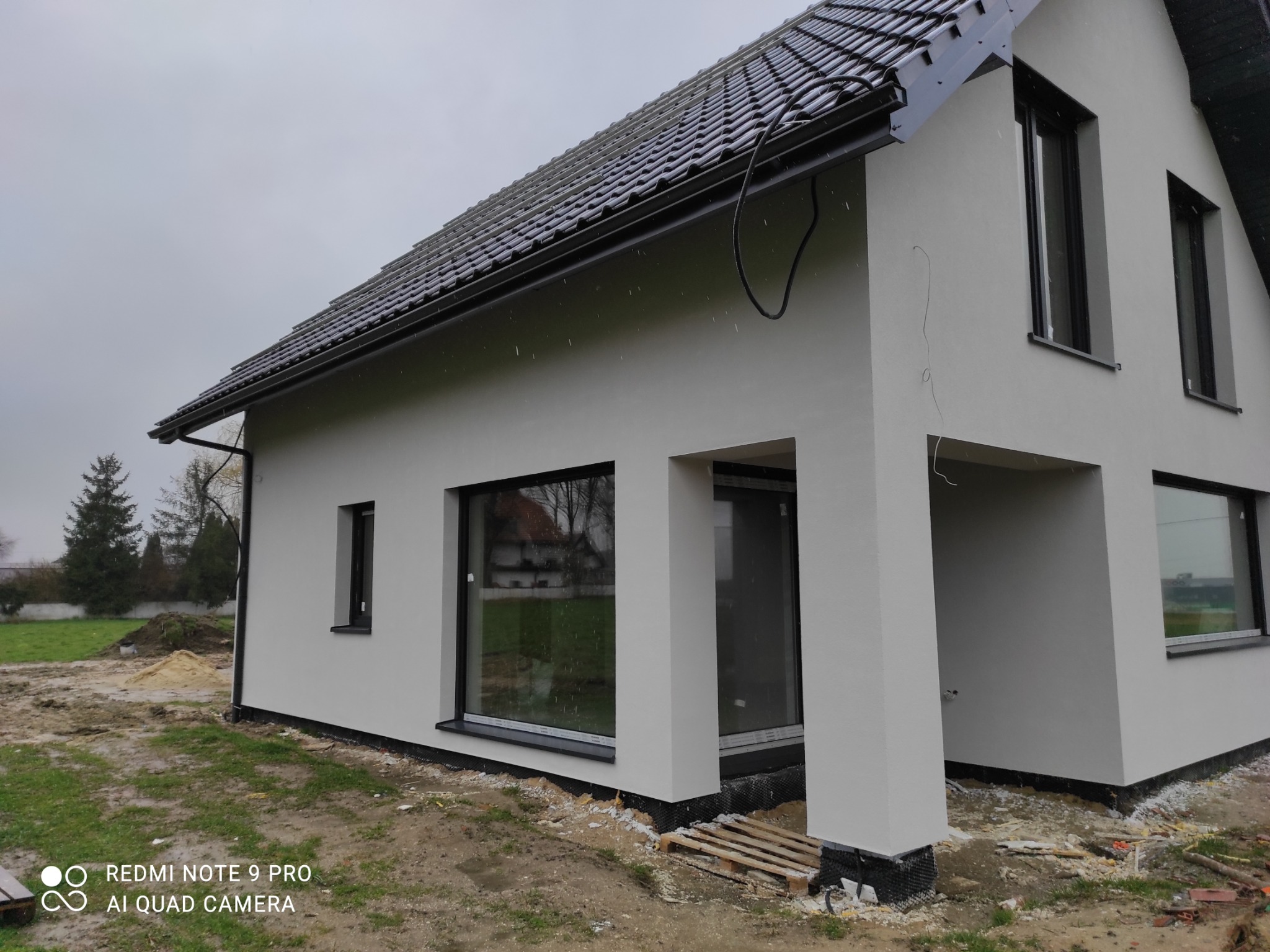 Nowoczesny dom jednorodzinny z szarą elewacją i czarną stolarką okienną, widoczne ślady prac wykończeniowych wokół budynku, pochmurne niebo.
