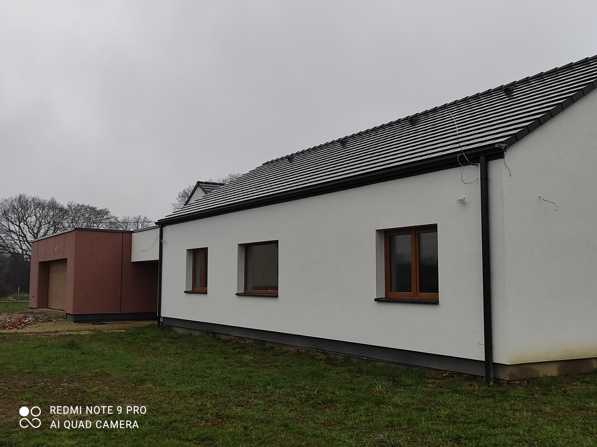 Nowoczesny dom jednorodzinny z białą elewacją i brązowymi oknami, widoczny garaż z elewacją w odcieniach brązu, trawnik z przodu, pochmurne niebo w tle.