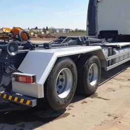 Firma usługowa Kacper Juskowiak - Tył ciężarówki z platformą hakową, widoczne elementy hydrauliczne i zabezpieczenia odblaskowe, teren budowy w tle.