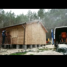 Firma usługowa Kacper Juskowiak - Malowanie drewnianego domku letniskowego na betonowych bloczkach, widoczny pracownik w trakcie nanoszenia impregnatu, w tle las i samochód dostawczy z urządzeniem do malowania.