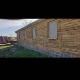 Firma usługowa Kacper Juskowiak - Drewniana elewacja budynku z zabezpieczonymi oknami przed malowaniem, widoczny fragment kamiennej podmurówki i trawnika, w tle czerwona laweta.