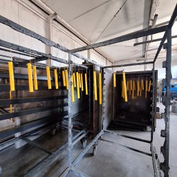 Firma usługowa Kacper Juskowiak - Żółte, cylindryczne elementy zawieszone na hakach wewnątrz metalowej konstrukcji, prawdopodobnie przygotowane do procesu nakładania powłoki; widoczny fragment pieca.