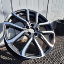 Felga aluminiowa samochodowa, pomalowana proszkowo na kolor grafitowy z srebrnymi akcentami, leży na gumowej wycieraczce na tle szarej ściany z paneli.