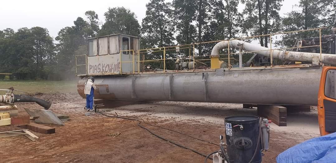 Pracownik w kombinezonie ochronnym czyści strumieniowo dużą, metalową konstrukcję pływającą z napisem 'PIASKOWANIE' na tle drzew i łąki, z widocznym sprzętem do piaskowania.