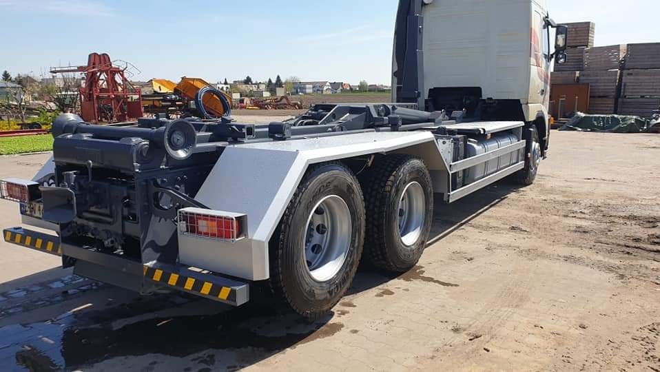 Tył ciężarówki z platformą hakową, widoczne elementy hydrauliczne i zabezpieczenia odblaskowe, teren budowy w tle.
