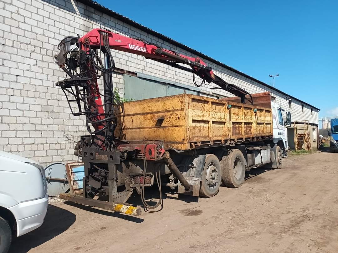 Ciężarówka z żółtą, rdzawą skrzynią ładunkową i czerwonym dźwigiem VKRANE, zaparkowana na placu przed budynkiem z białej cegły.