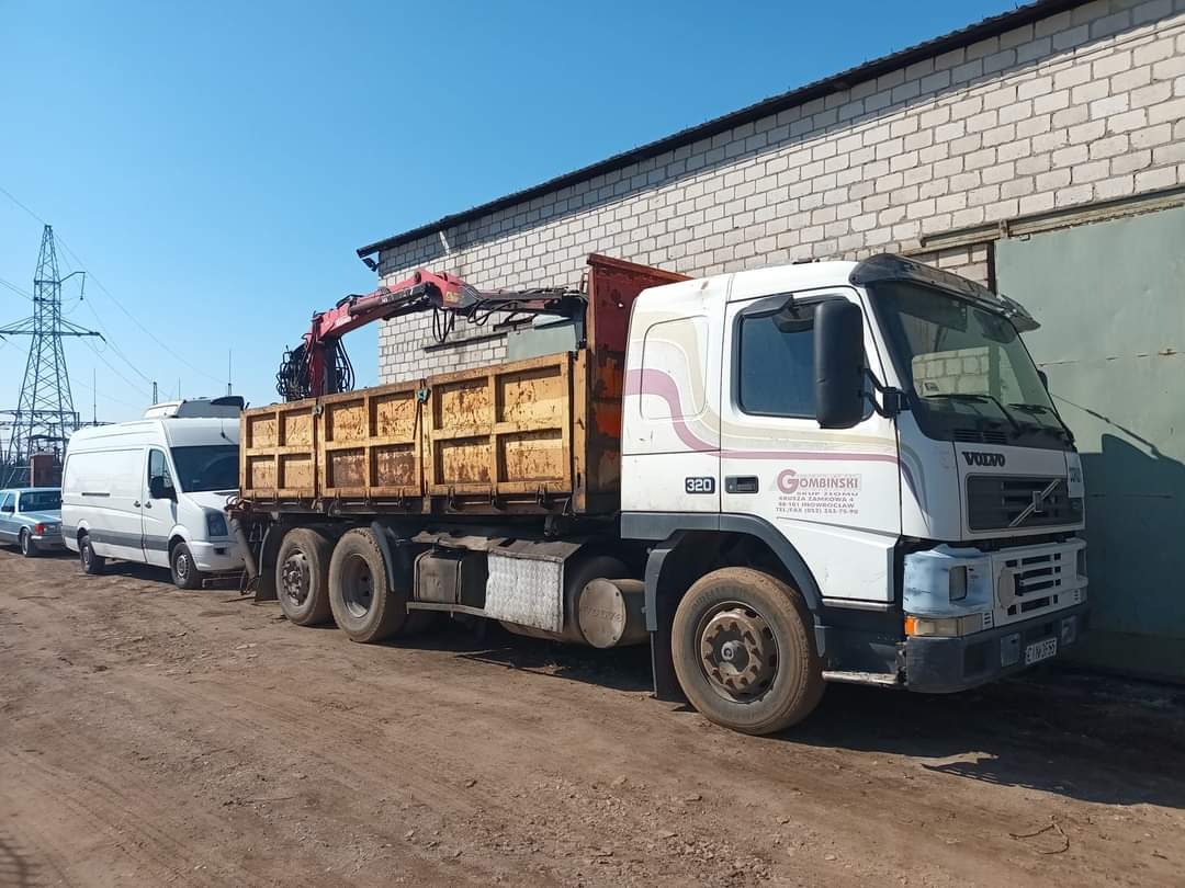 Ciężarówka Volvo z hydraulicznym dźwigiem załadowcza z otwartą skrzynią na tle murowanego budynku i słupów wysokiego napięcia.