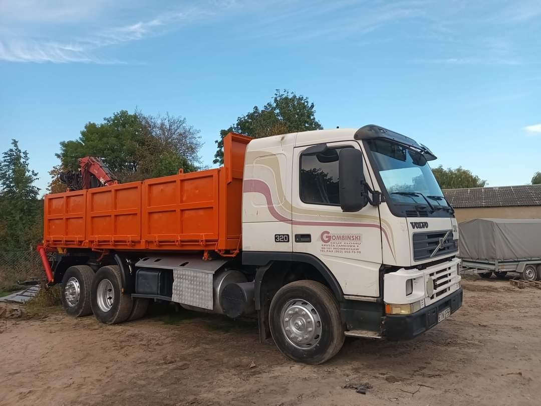 Pomarańczowa wywrotka Volvo z żurawiem HDS na tle błękitnego nieba i zieleni drzew, zaparkowana na piaszczystym gruncie.