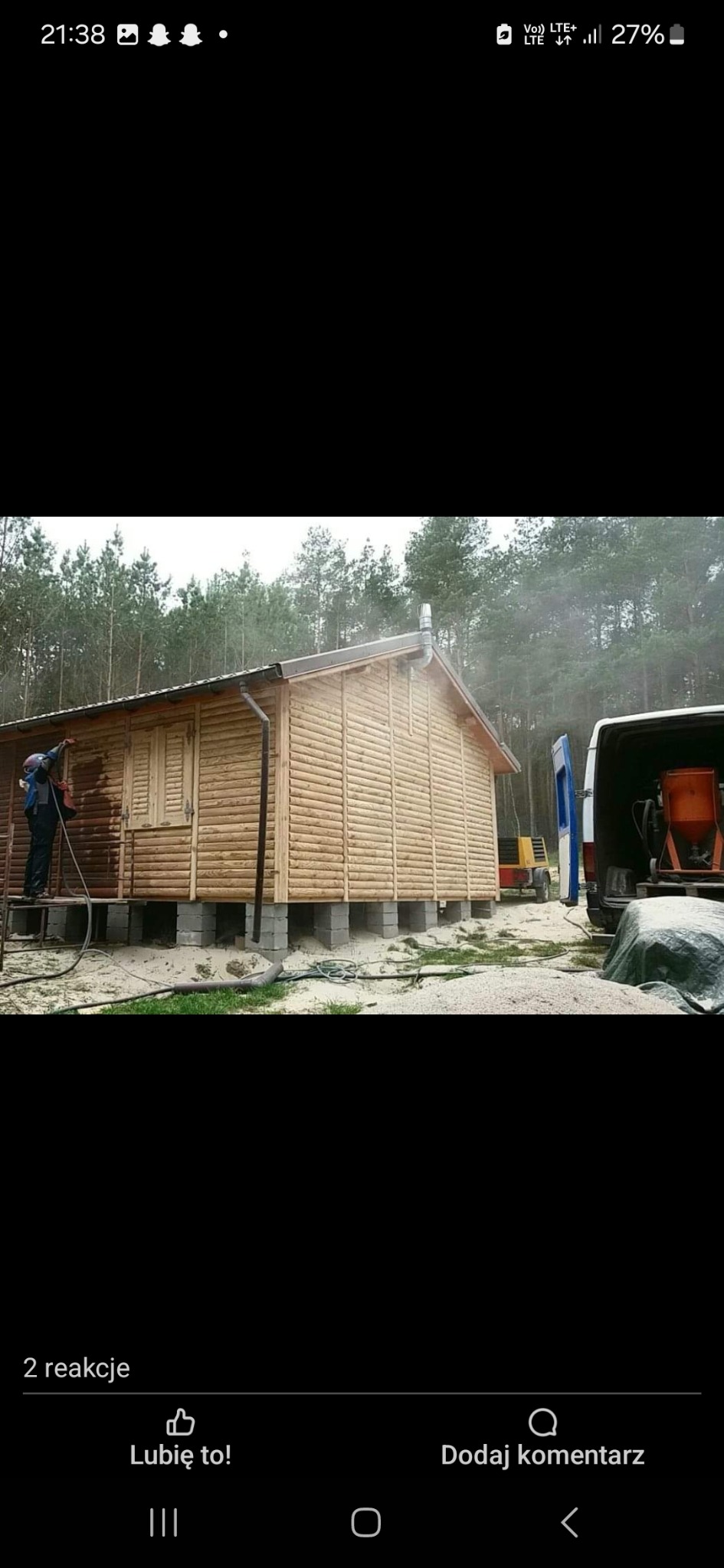 Malowanie drewnianego domku letniskowego na betonowych bloczkach, widoczny pracownik w trakcie nanoszenia impregnatu, w tle las i samochód dostawczy z urządzeniem do malowania.